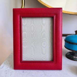 Op de afbeelding: Een rechthoekige rode fotolijst met een textuur die op leer lijkt. De lijst heeft een kleinere binnenrand en een lichtgekleurde achtergrond met patronen. De lijst staat op een marmeren oppervlak.