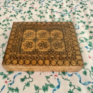 May include: A rectangular wooden box featuring a dark brown pyrography design on the lid. The center displays a grid of six flower motifs, surrounded by a border of repeating circles and intricate line patterns etched into the light wood surface.