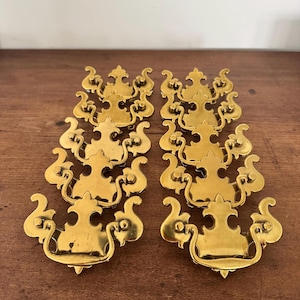 Könnte beinhalten: Acht verzierte, goldfarbene Schubladengriffe, angeordnet auf einer Holzoberfläche. Jeder Griff weist ein dekoratives Design mit geschwungenen Elementen und einem Griff auf. Die Beschläge sind wahrscheinlich für die Möbelrestaurierung gedacht.