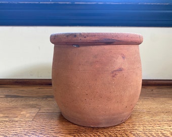 Puede incluir: Una maceta de terracota con forma redondeada y un borde ligeramente acampanado. La maceta tiene un aspecto rústico y envejecido, con un color marrón moteado e imperfecciones visibles.