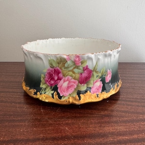 Puede incluir: Un cuenco redondo de porcelana con un fondo degradado que va del verde oscuro al blanco. El exterior está decorado con racimos de rosas rosadas y hojas verdes, con un borde y una base festoneados pintados en color oro.