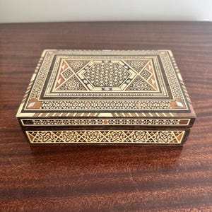 May include: A rectangular wooden box featuring intricate geometric marquetry inlay on the lid and sides. The design incorporates light and dark wood tones with small mother-of-pearl accents arranged in complex star and diamond patterns.