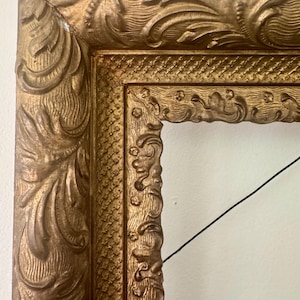 May include: Ornate gold picture frame with detailed carvings and a textured surface. The frame has a rectangular opening and is set against a white background. The frame's design features scrolling patterns and decorative elements.