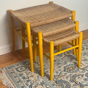 May include: A set of three nesting tables. Each table has a woven, light brown top and bright yellow legs. The tables are of varying sizes, designed to stack together for storage. The tables are made of wood.