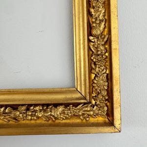 May include: A gold-colored picture frame with ornate floral and leaf detailing. The frame has a rectangular shape with a decorative border of sculpted flowers and foliage. The frame is made of a shiny, metallic material.