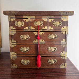 May include: A small, wooden chest of drawers with brass hardware and decorative corner accents. The chest features multiple drawers with ornate pulls and a red tassel. The wood has a rich, dark brown finish.