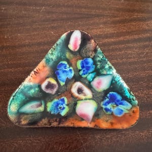 May include: A triangular enameled brooch featuring a vibrant abstract pattern. The surface displays a blend of deep green, orange, and black tones, accented by scattered, raised, glossy shapes in shades of bright blue, white, and soft pink.