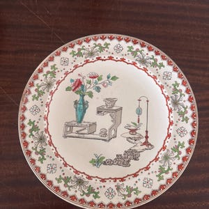 May include: A round ceramic plate featuring a cream-colored center with a detailed illustration of a teal vase, a wooden stand, and a tall lamp. The rim is decorated with a repeating pattern of red, green, and white floral and geometric motifs.