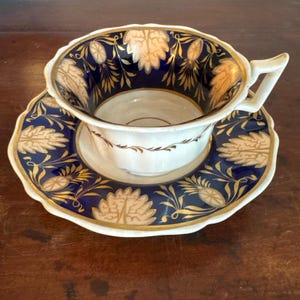 May include: A white teacup and saucer set with a navy blue and gold floral design. The cup has a scalloped rim and a curved handle. The saucer mirrors the cup's design, creating a cohesive and elegant aesthetic.