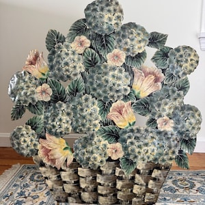 May include: A metal floral basket sculpture featuring blue-green hydrangeas, pink and yellow flowers, and green leaves. The basket has a woven design and is painted in neutral tones. The sculpture is placed on a patterned rug.