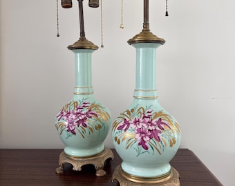 May include: A pair of antique porcelain table lamps with a light blue base and gold accents. The lamps feature hand-painted floral designs in shades of pink and purple.