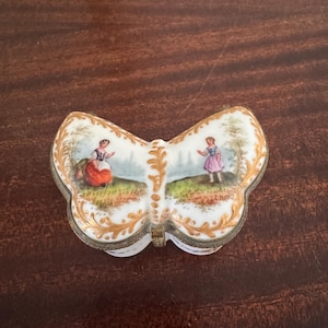 May include: A butterfly-shaped porcelain trinket box with a white base, gold filigree trim, and hand-painted scenes of two figures in a landscape. The hinged lid features a metal clasp and detailed illustrations of people in colourful clothing.
