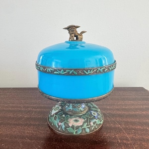 Puede incluir: Un tarro decorativo azul turquesa con tapa, con una figura de perro de color bronce en la parte superior. El tarro tiene una base con estampado floral y una banda decorativa. El tarro se utiliza probablemente para guardar objetos pequeños.