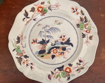 Puede incluir: Un plato de cerámica blanca con borde festoneado, con una escena central de paisaje y un borde floral. El diseño incluye flores y follaje de color naranja, azul, verde y rosa. El plato está sobre una superficie de madera oscura.