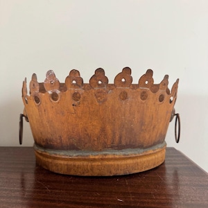 Puede incluir: Un cuenco de metal vintage en forma de corona con un aspecto envejecido y desgastado. El cuenco es de color marrón óxido cálido con un borde decorativo festoneado. Cuenta con dos asas circulares pequeñas y descansa sobre una base de madera.