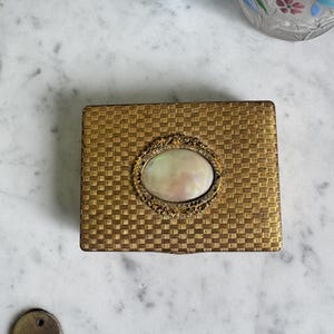 May include: A rectangular, gold-toned vintage box with a woven pattern. An oval, iridescent mother-of-pearl inlay is set in a decorative frame on the lid. The box is likely for jewelry or small keepsakes.