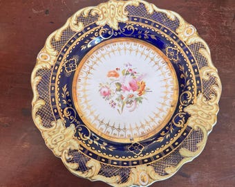Puede incluir: Un plato decorativo con un borde azul y dorado y un centro blanco con un diseño floral. El plato tiene un borde festoneado y detalles dorados intrincados.