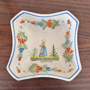 May include: A square, off-white ceramic dish with a scalloped edge and a blue border. The dish is decorated with floral patterns in green, orange, and blue. A central illustration depicts a woman in traditional attire. The text "HR Quimper" is written below the illustration.
