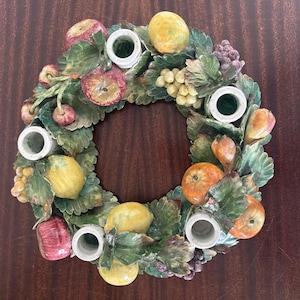 May include: A circular ceramic candle holder featuring a decorative arrangement of sculpted green leaves and various fruits including red apples, yellow lemons, oranges, and clusters of purple and green grapes, with five white cylindrical candle cups.