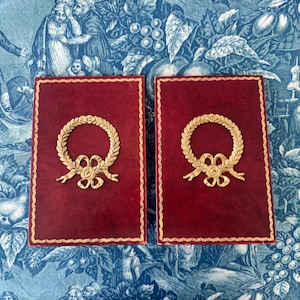 May include: Two rectangular books with deep red velvet covers, each featuring a centered gold-toned metal laurel wreath emblem with a ribbon detail at the bottom. The covers are bordered by a thin, decorative gold-toned filigree line pattern.