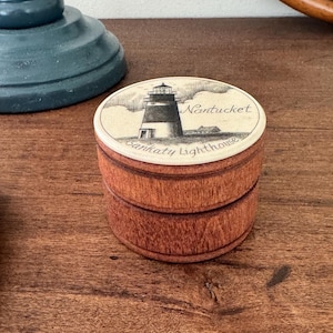 Puede incluir: Una pequeña caja redonda de madera con tapa. La tapa presenta una ilustración en blanco y negro de un faro con las palabras "Nantucket" y "Sankaty Lighthouse" en escritura. La caja es de madera marrón rojiza.
