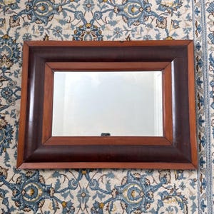 Puede incluir: Espejo rectangular con marco de madera. El marco tiene un borde exterior marrón oscuro y un borde interior marrón más claro. El espejo es rectangular y refleja el entorno. Es un artículo decorativo.