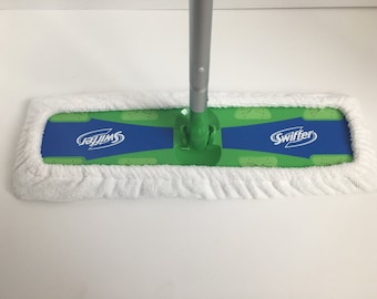 May include: A white Swiffer Sweeper mop head with a green and blue cleaning pad. The pad has the word "Swiffer" printed on it in white.