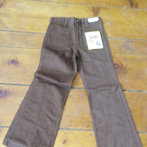 Puede incluir: Vaqueros marrones de pierna ancha con una etiqueta de Wrangler. Los vaqueros tienen una abertura de pierna ancha y un ajuste recto a través del muslo.