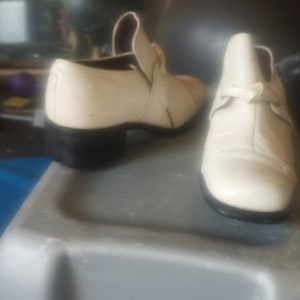 Könnte beinhalten: Ein Paar cremefarbene Lederschuhe mit schwarzen Sohlen. Die Schuhe haben eine spitze Zehenpartie und einen niedrigen Absatz.