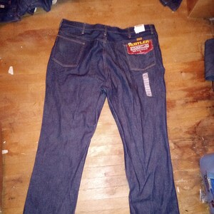 Könnte beinhalten: Eine blaue Jeans mit einem braunen Leder-Patch auf der Gesäßtasche, auf dem "Rustler" in gelben Buchstaben steht. Die Jeans hat einen geraden Schnitt und eine lockere Passform.