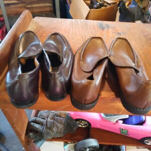 May include: Four pairs of brown leather loafers on a wooden shelf. The loafers have a slip-on design and are arranged in two pairs.