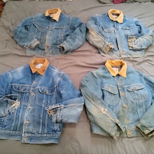 May include: Four vintage denim jackets with light blue washes and tan corduroy collars are displayed. The jackets have button closures, pockets, and show signs of wear, including distressed areas on the sleeves and body. The jackets are laid out on a gray surface.