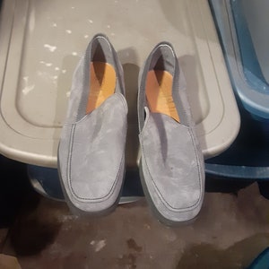 May include: A pair of gray suede slip-on shoes with a dark gray trim around the edges.