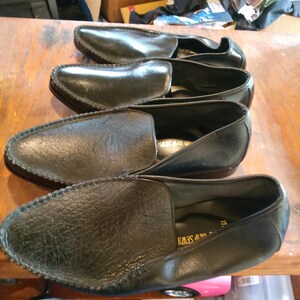 May include: Three pairs of black leather loafers with a decorative stitch around the edge. The loafers have a pointed toe and a slip-on design.