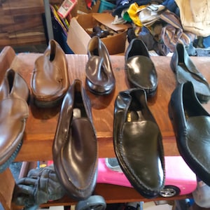 May include: Seven pairs of leather slip-on shoes in various shades of brown and black. The shoes are arranged on a wooden surface.