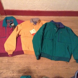 Op de afbeelding: Drie jassen in verschillende kleuren: een bordeauxrode jas met een turquoise kraag, een mosterdgele jas met een grijze kraag en een groene jas met een blauwe kraag.
