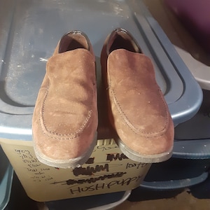 May include: A pair of brown suede Hush Puppies slip-on shoes. The shoes are sitting on top of a blue plastic storage bin.