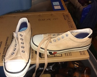 May include: Two pairs of beige canvas sneakers with white soles and black trim. One pair has blue trim and the other has red trim. The sneakers are sitting on top of a cardboard box with writing on it. The box is sitting on top of a brown plastic bucket filled with other shoes.