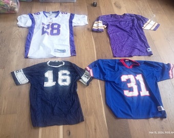 May include: Four American football jerseys are laid out on a wooden surface. The jerseys are white, purple, navy blue, and royal blue. Each jersey has a number on the front and back. The jerseys are short-sleeved.