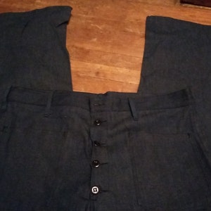 Può includere: Un paio di pantaloni di jeans blu scuro con chiusura a bottoni e cinque tasche. I pantaloni hanno una gamba larga e sono realizzati in un tessuto spesso e resistente.