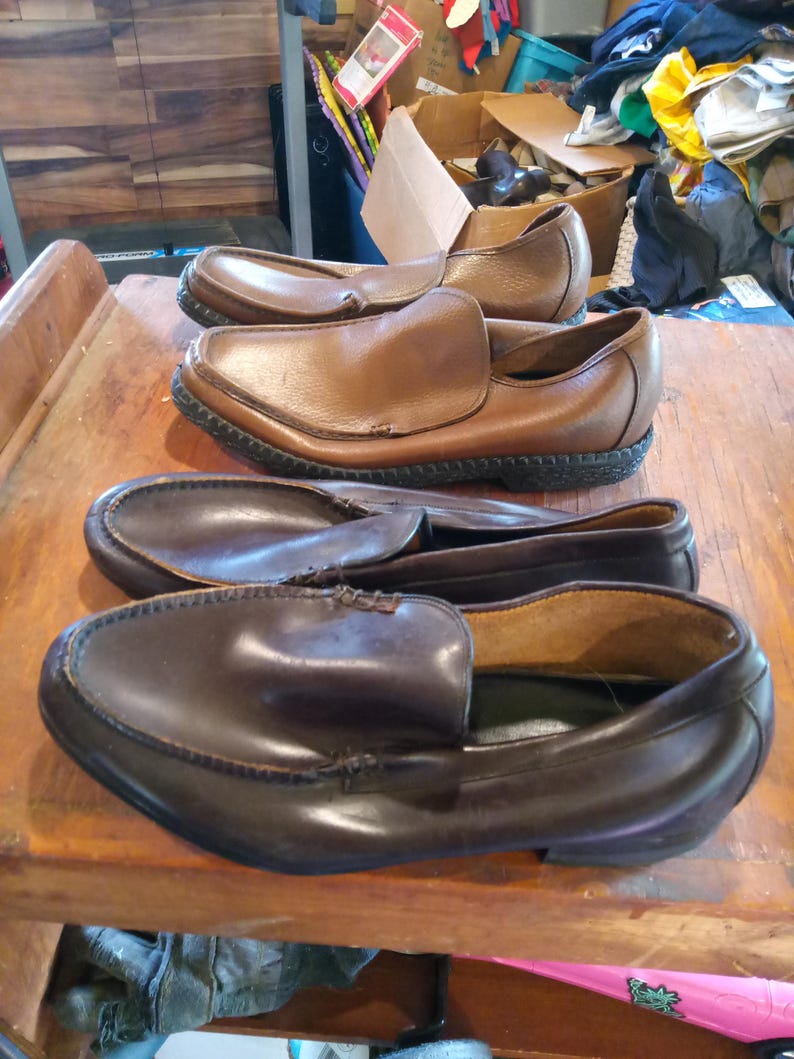 May include: Three pairs of leather slip-on shoes. The shoes are brown and dark brown in color. The shoes have a stitched seam around the edge of the sole.
