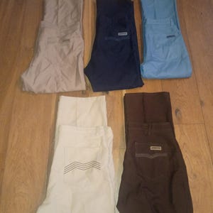 May include: Six pairs of pants in various colors are displayed on a wooden surface. The colors include beige, navy blue, light blue, off-white, and brown. Each pair has a pocket design and a small rectangular label.