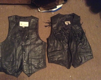 May include: Two black leather vests. One vest has fringe detailing and the other has a zipped pocket. Both vests have a label with the word "Genuine" on them.