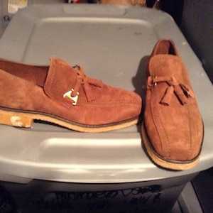 May include: A pair of brown suede tassel loafers with gold accents. The loafers have a leather sole and a slightly pointed toe.