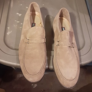 May include: A pair of light brown suede loafers with a black strap across the top. The loafers have a brown leather sole and a slightly pointed toe.