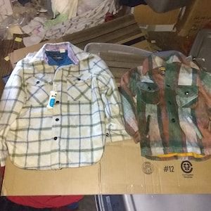 May include: Two plaid flannel shirts, one white and green with black checks, the other with brown, green, and orange stripes. Both shirts have button closures and pockets.