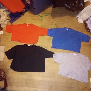 Peut inclure: Quatre t-shirts coupés à différentes couleurs : rouge, bleu, noir et gris. Les t-shirts sont disposés sur un sol en bois.