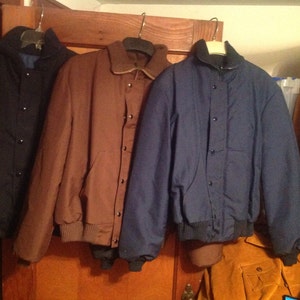 May include: Three jackets hanging on hangers. The jacket on the left is black, the middle jacket is brown, and the jacket on the right is navy blue.
