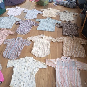 May include: A collection of short-sleeved button-down shirts in various colors and patterns, including floral, striped, and solid designs. The shirts are laid out on a wooden floor, showcasing a variety of styles and fabrics.