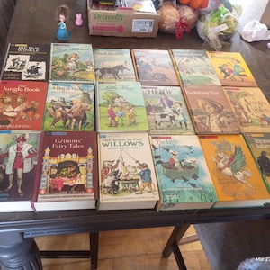 Op de afbeelding: Een verzameling vintage kinderboeken met kleurrijke covers, waaronder titels als "The Jungle Book", "Grimms' Fairy Tales" en "Treasure Island". De boeken zijn gerangschikt op een donkere houten tafel.